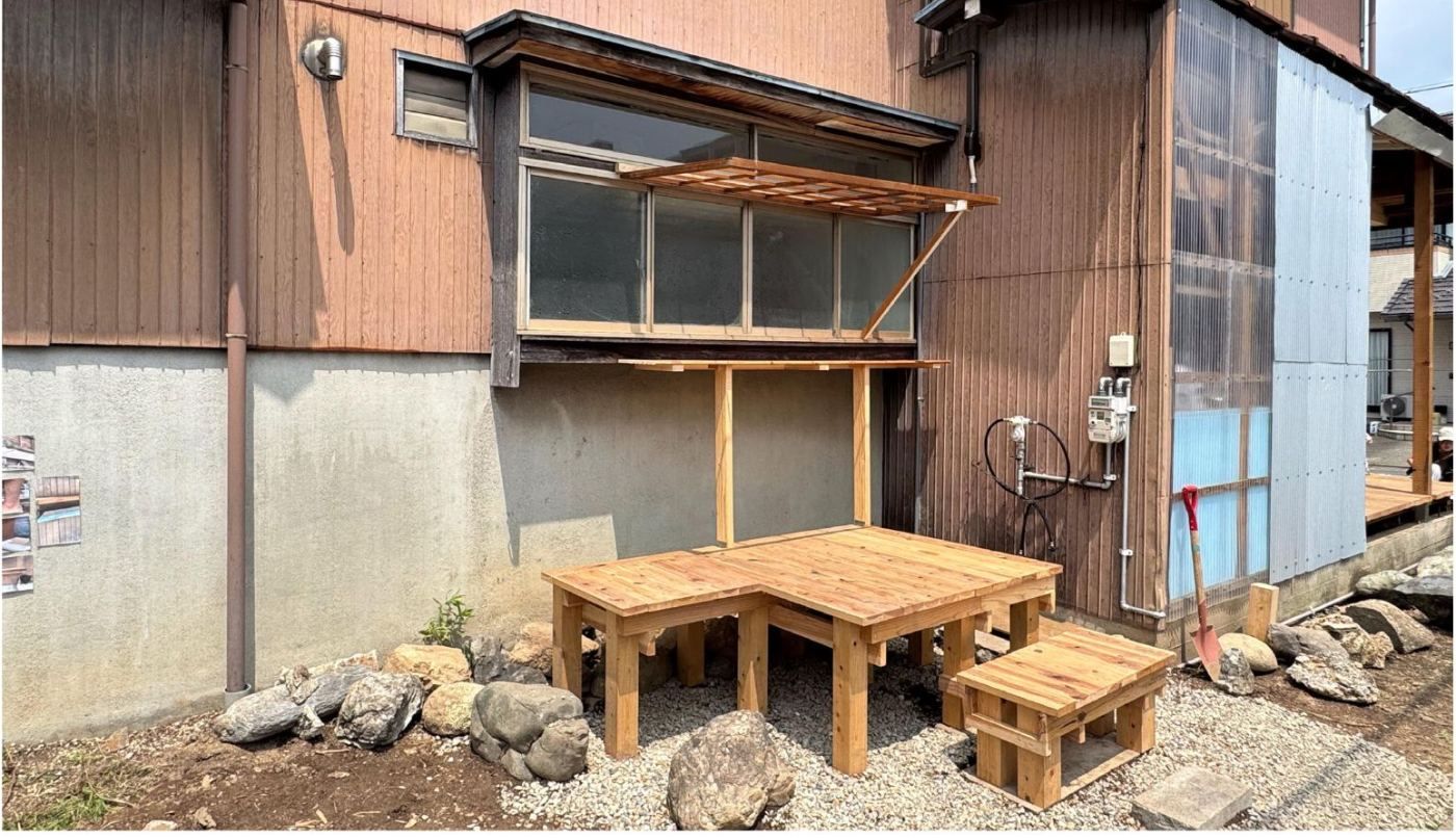A low timber deck with a built-in table and bench outside a timber-clad house in Gifu, ringed by rocks